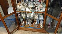 Wide view showing glass cabinet with assorted porcelain figurines including angels and cherubs, decorative plates, clock under glass dome, and other decorative items.