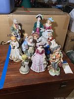 Overview of 11 assorted porcelain figurines with various period costumes and poses, displayed on wooden surface against cardboard boxes