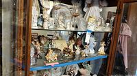 Photo of curio cabinet shelf with multiple Precious Moments porcelain figurines including children and angels, some holding animals or hearts, and various small decorative items.