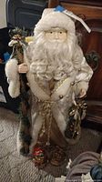 Front view of Santa doll showing white glittery coat with white faux fur trim and bells, along with staff and gift sack