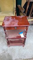 Front view of vintage wood side table showing top surface, drawer with metal pull, open shelf, and cabinet door below. Table has reddish-brown finish with visible dust and wear.
