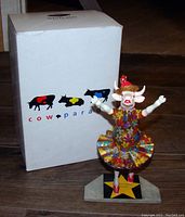 CowParade Dancing Diva figurine with original box pictured on a wooden floor, showing the colorful cow in a tutu with arms raised, on a star base with 'Dancing Diva' text.