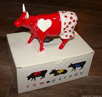 Photo of ceramic cow figurine Moocho Amor from CowParade, showing full figure on top of box with CowParade logo and colorful cow silhouettes