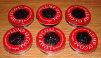 Six round red-lid tins visible with black lump of coal inside, tops readable with white text 'Lump O'Coal Because you've been very naughty' on wooden surface.