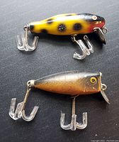 Two vintage wooden fishing lures displayed on black surface. Bumble Bee lure is yellow with black spots and red on head, with plastic treble hooks and metal lip. Jitterbug lure is brown with yellow eye and plastic treble hooks attached.
