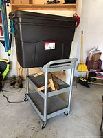 Full view of gray plastic utility cart with one Rubbermaid tote on top and miscellaneous items on lower shelves in garage.