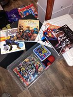 Photo showing multiple Marvel and DC comic-related books including Marvel Encyclopedia, DC Comics Encyclopedia, Spider-Man, and X-Men books with various items in a plastic bin.