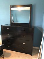 Front view of black dresser with mirror attached, showing six drawers with silver handles and the mirror reflecting the room.