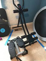 Photo showing the Sunny Health & Fitness stepper with textured foot pedals and handles placed on a carpeted floor next to other fitness equipment