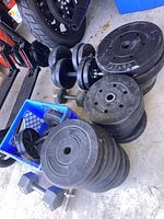 Photo showing free weight plates stacked on floor, two adjustable dumbbell bars with collars, and two fixed rubber hex dumbbells visible near a blue crate and a motorcycle wheel.