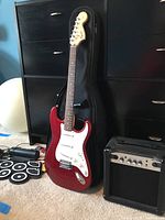Red Fender Squier Strat electric guitar leaning against a black dresser with soft case behind and Marshall amplifier to the right