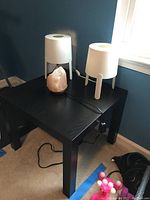 Black square wooden side table with three table lamps on top positioned near a window: one pink Himalayan salt lamp and two white tripod-style lamps.