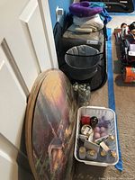 Lot contents including folded bed linens, neck pillows, black mesh laundry net wastebasket, glass vase with shells, and white basket with bath products arranged on carpet against white door.