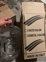 Glass carboy held next to its original brown cardboard box labeled '3 Water Gallon' and 'Cosmetal 3 Gallon', showing size and packaging details.