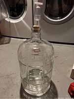 Full view of the 3 gallon clear glass carboy with rubber stopper and plastic air lock attached, standing on a concrete floor.