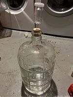 Front view of 3 gallon clear glass carboy with rubber stopper and transparent air lock attached, sitting on floor with washer and dryer in background.