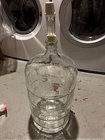 Full view of 6 gallon glass carboy with plastic stopper and clear airlock attached on top, shown on gray floor.