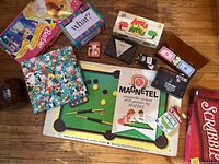 Top view of assorted games spread on the floor showing colorful boxes including Scrabble, Apples to Apples, What?, Magnetel by Mattel, Exploding Kittens NSFW, and game dice.