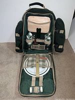 Full view of backpack opened displaying picnic plates, glasses with green checkered covers, salt and pepper shakers, and empty utensil holders.