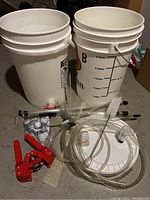 Two fermentation buckets, one marked with gallon measurements and multiple clear siphon tubes coiled on a white bucket lid, with red bottle capper tool and metal caps on gray floor.
