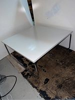 Full view of the square coffee table with shiny white top and tubular chrome metal legs, placed on a worn floor with plain wall background.