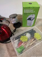 Photo showing red electric kettle, mini cast iron skillet, burger maker/stacker, and a container with various kitchen utensils, plus the green and white Everyday Essentials popcorn maker box.