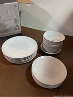 Set of white porcelain large plates stacked next to small plates and bowls. Box in background does not match set.