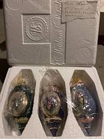 Three porcelain ornaments wrapped in plastic inside a Styrofoam box with embossed branding and handwritten label.
