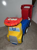 Front angled view of Little Tikes push truck showing yellow front, blue seat, red handle, and smiling cartoon eyes on windshield.