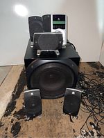 View of five Logitech satellite speakers, central channel speaker, subwoofer, and control console with cables on side placed on wooden floor.