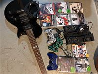 Photo showing the black electric guitar with cable, 3 PlayStation controllers including camo and lightning designs, 2 PS3 Move charging bases, 2 charging cables, and 9 PS3 game cases.