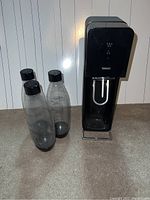 Full view of SodaStream machine and three bottles on floor against white panel wall.
