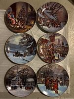 Top view of six decorative porcelain plates featuring vintage Christmas scenes from the W S George Scenes of Christmas Past series, showing detailed painted winter holiday imagery.