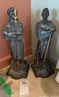 Pair of metal sculptures of men standing on ornate bases placed on carpet against wall.