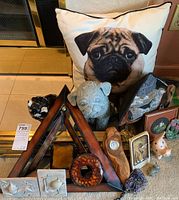 Lot arranged with pillow featuring pug face behind resin dog statue and wooden thermometer piece, including metal wall art and small decorative objects like ceramic animals and framed items