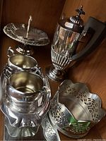 Photo showing silver plated coffee pot, pedestal candy dish, creamer, sugar bowl, small handled dish, and tray on wooden surface.