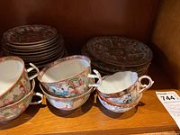 Tea cups stacked with matching saucers showing detailed hand painted figures and floral motifs in a cabinet