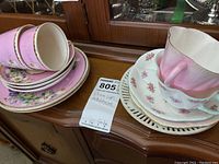 Six pink floral-patterned teacups and saucers with daisy designs stacked, showing delicate gold trim and detailed floral arrangements.