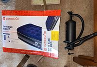 Outbound Twin Size Air Bed in box next to black hand pump on carpeted floor.