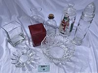 Full view of all glassware items arranged on white fabric background, showing two square vases (one clear one red), scalloped bowl, jars with lids including one decorated jar, bottle with cork, and star-shaped dish.