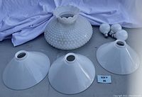 Photo showing one white milk glass Fenton light shade with hobnail pattern, three white metal cone light shades, and three light bulbs arranged on a grey surface with a purple cloth background.