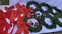 Six green artificial wreaths, red bows and assorted pinecones and picks laid out on white fabric.
