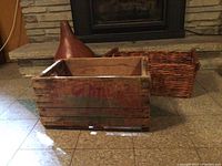 Wooden crate with distressed red painted lettering and a woven rectangular wicker basket behind it, placed on tiled floor in front of fireplace.