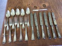 Photo showing 6 forks, 6 spoons, 7 knives with patina and tarnish, and one small spreader knife arranged on wooden surface.