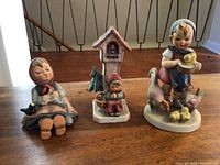 Three Goebel West Germany porcelain figurines displayed on wood surface: girl with chicks and hen, child kneeling at birdhouse, girl sitting with bird