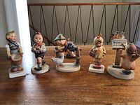 Full view of all five Hummel figurines in a row on wooden surface showing their front details.