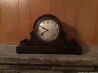 Front view of the dark wooden mantel clock placed on a stone surface against a wood-paneled wall.