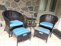 Photo of full patio set including two wicker barrel chairs with blue cushions, two matching ottomans, and a round side table with glass top on an outdoor stone patio against stone wall and windows.