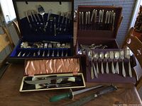 Multiple flatware sets displayed outside their open cases showing knives, forks, spoons, and carving set with cream handles along with two loose carving knives.