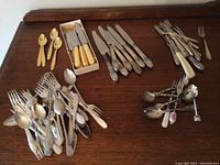 Photo of assorted forks, spoons, knives including some with cream-colored handles, all laid out on wooden table.
